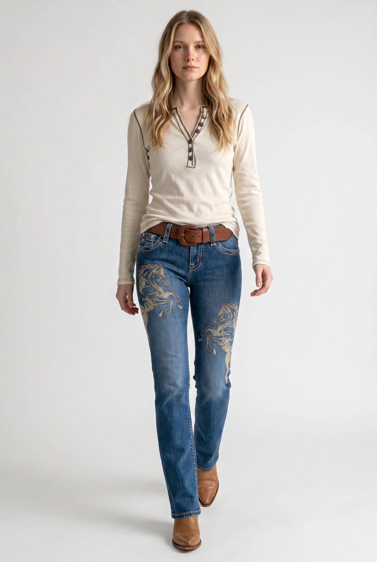 A woman modeling blue jeans with gold floral embroidery on the thighs, paired with a cream long-sleeve henley top, a brown belt, and brown boots, against a light gray studio background.