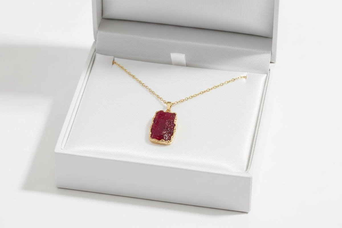 A gold necklace with a rectangular red gemstone pendant, displayed inside an open white jewelry box on a clean white background.