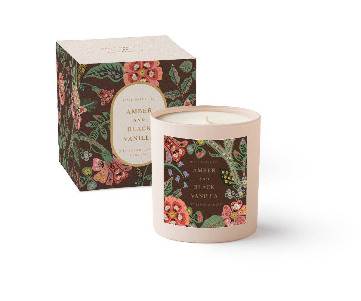 Rifle Paper Company Gracie Amber and Black Vanilla 9 oz glass candle with floral design and black lid on a wooden surface.